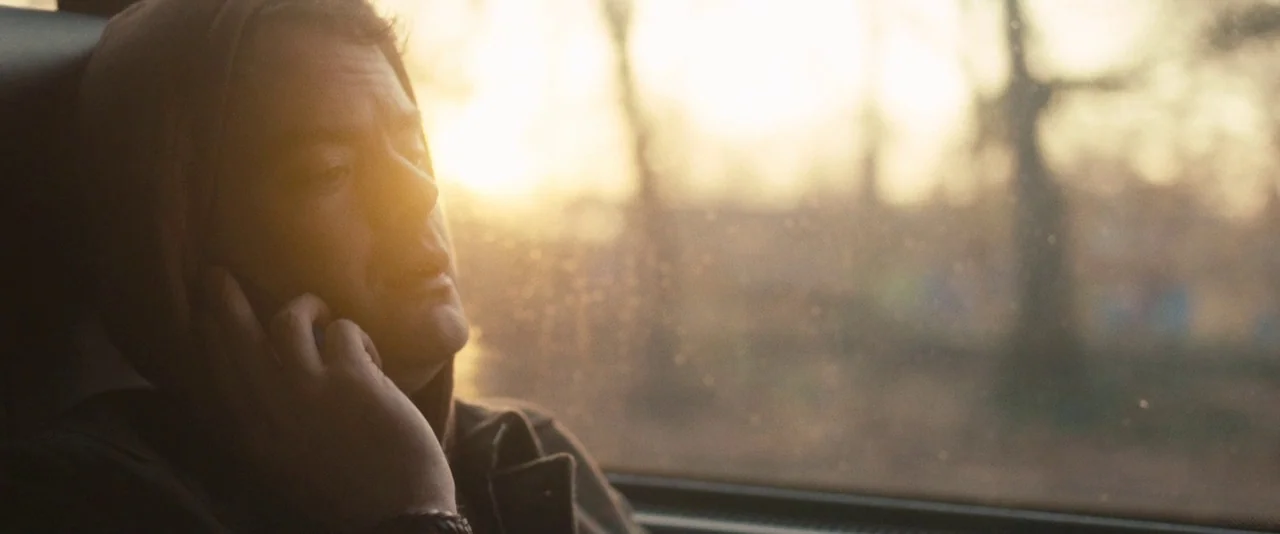 The Skeleton Twins (2014) movie still: closeup — A person sits beside a window inside a train during the late afternoon. The warm sun illuminates…