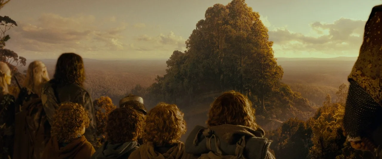 A group of travelers stands on a high mountain ridge looking out over a valley. A vast forest stretches toward the horizon under a soft golden sunset sky.