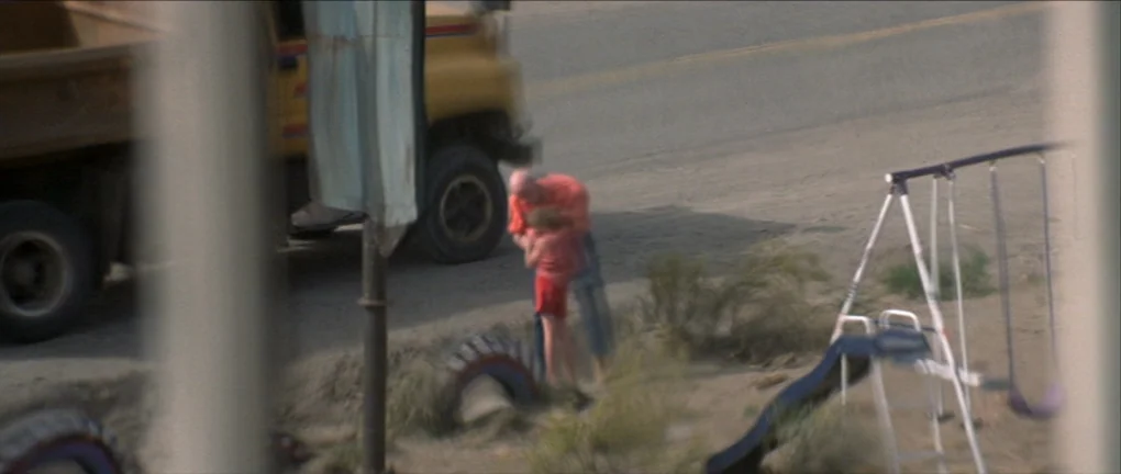 The Pledge (2001) movie still: wide — A dusty, unpaved roadside sits near a yellow truck and a child's swing set. An adult male and a small child…