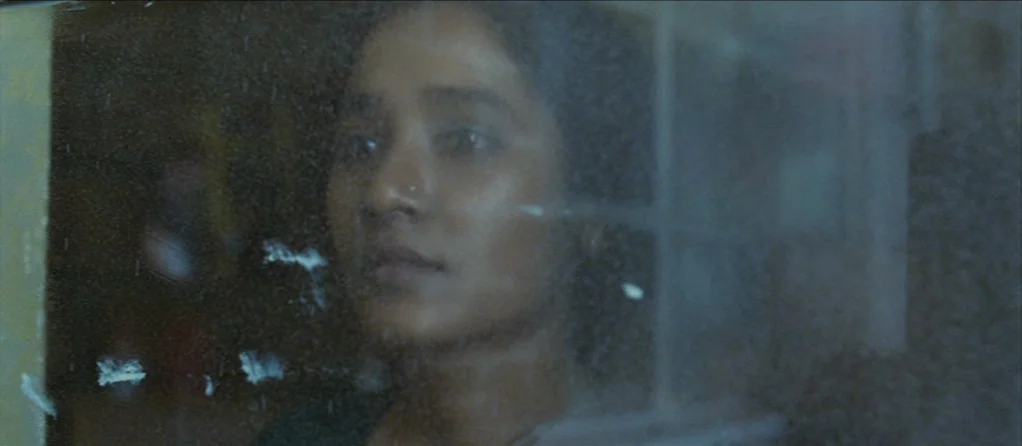 Brick Lane (2007) movie still: closeup — A person looks out through a transparent pane of glass covered in smudge marks. The subject is partially…