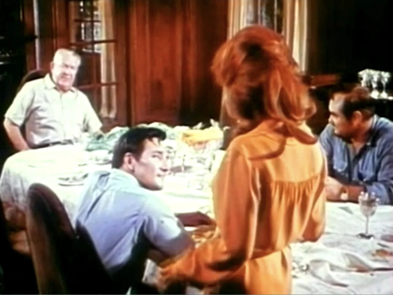 In the Year 2889 (1969) movie still: medium — A wood-paneled dining room contains a large table covered by a white tablecloth. A woman stands over a…