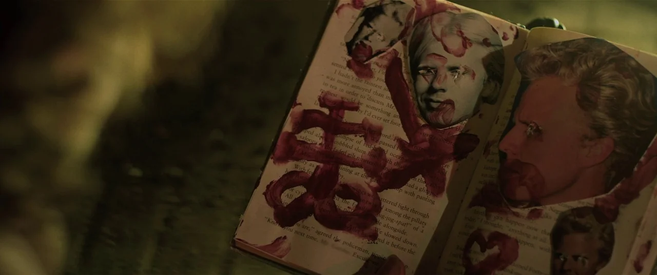 The Editor (2014) movie still: closeup — An open book resting in hands displays collaged photographs of faces marked with red ink. Thick, viscous…