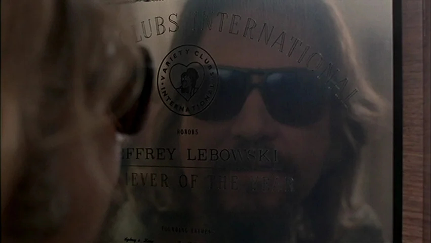 The Big Lebowski (1998) movie still: closeup — A framed award plaque hangs on a wooden wall displaying text about a recipient. A man's face is…
