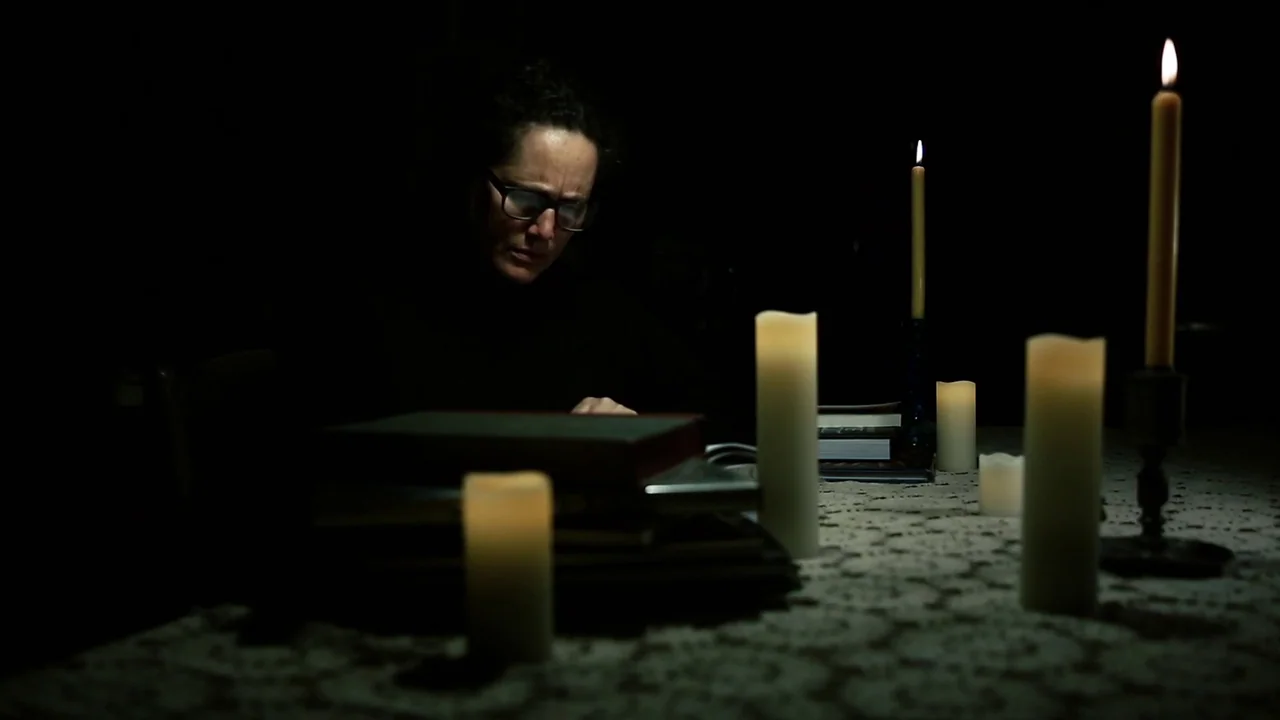 The Deeper You Dig (2019) movie still: medium — A dark dining room with a lace tablecloth and stacks of books. A woman sits at the table and reads…