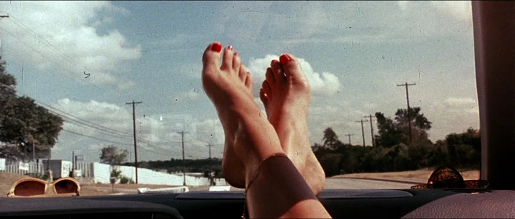 Death Proof movie still: closeup — A pair of feet with red toenail polish rests on a car dashboard. The view through the windshield shows a rural…