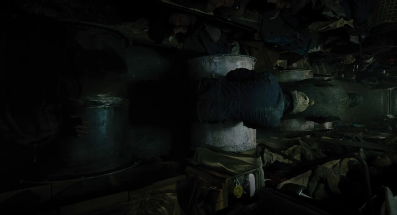 Snowpiercer (2013) movie still: wide — A dark, cramped, and metallic train compartment filled with people resting in low light. The passengers sit…