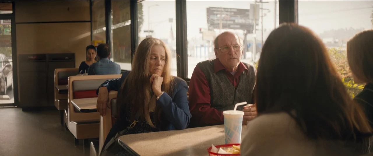 Kajillionaire (2020) movie still: medium — Two older people sit inside a diner booth facing an unseen person. Large windows behind them reveal a…