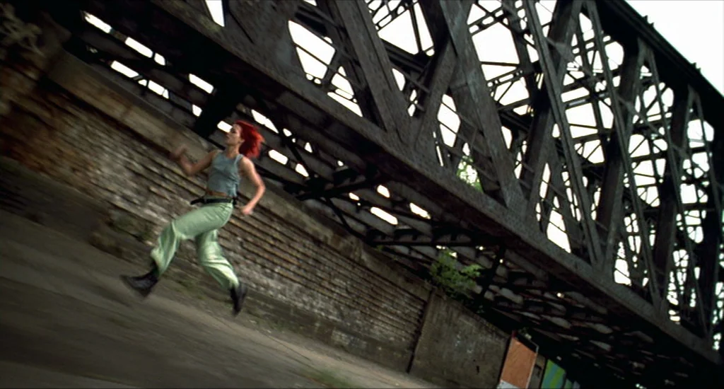 Run Lola Run (1998) movie still: wide — A rusted iron railway bridge spans over a paved ground area with brick walls. A young woman with red hair…