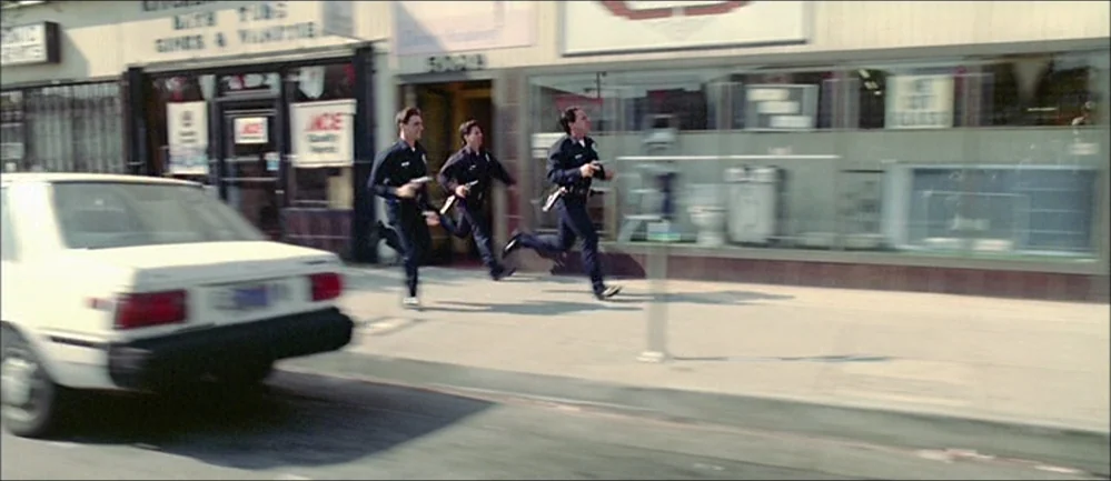 Reservoir Dogs (1992) movie still: wide — A sidewalk fronting commercial buildings features storefront windows and a parked white sedan. Three…