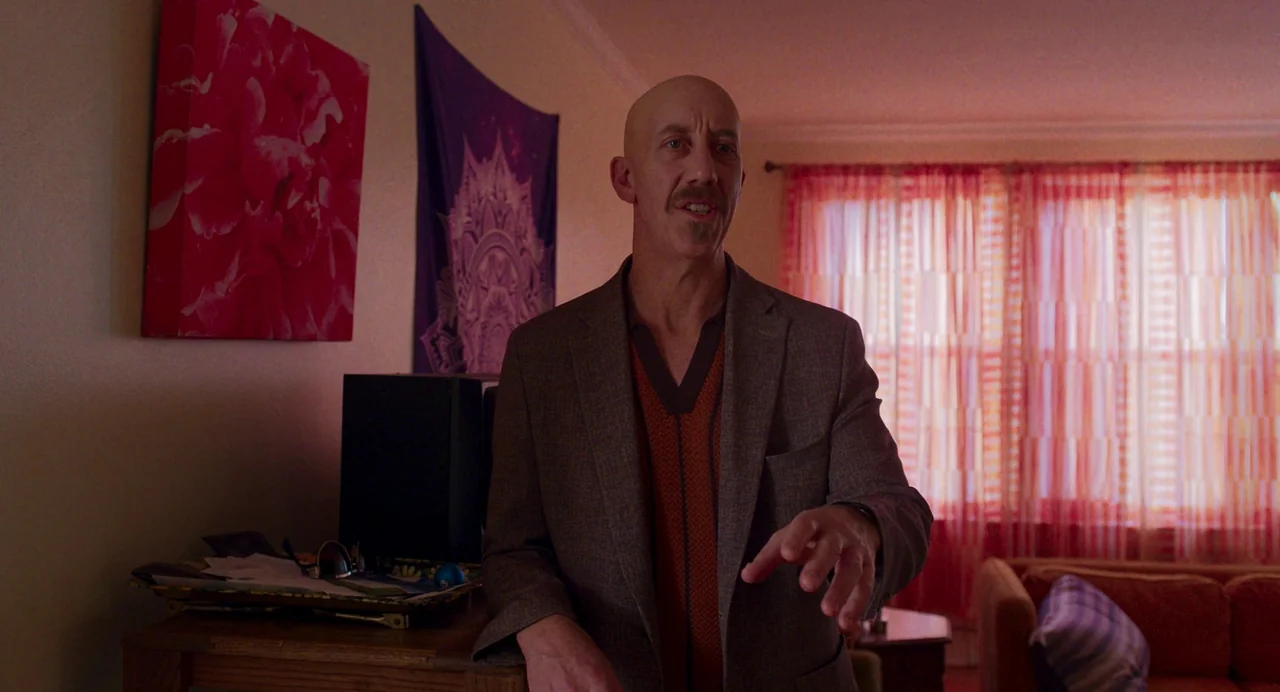 Drive-Away Dykes (2024) movie still: medium — A residential room features a purple tapestry and red wall art against the walls. A bald man wearing a…