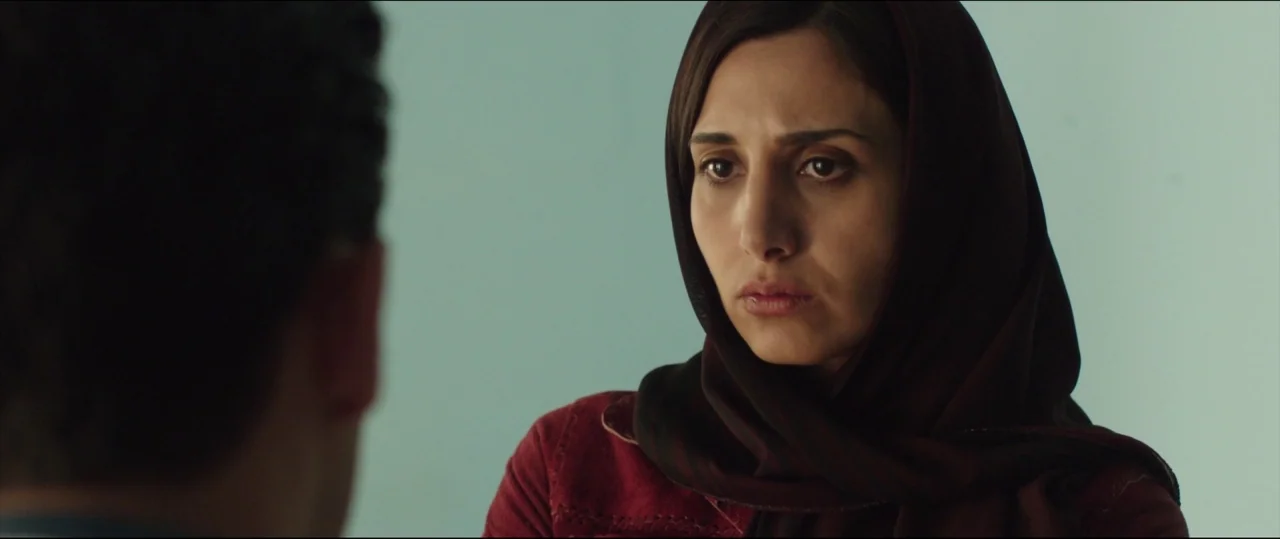 Under The Shadow (2016) movie still: medium — A woman wearing a headscarf sits in an interior space with plain teal walls. She maintains a fixed…
