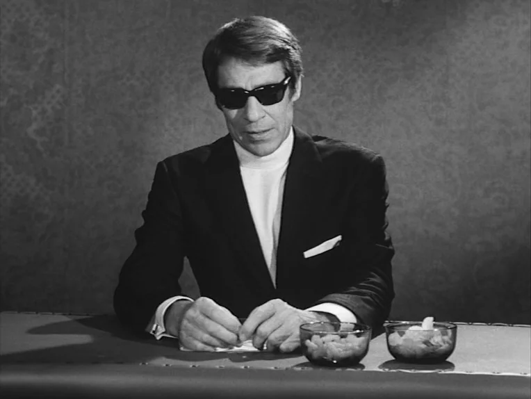 The Rite (1969) movie still: medium — A man sits before a solid-colored wall with faint textured wallpaper patterns. He rests his hands on a…