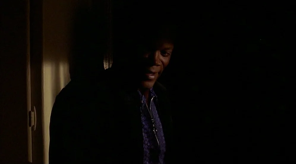 Jackie Brown movie still: medium — A dimly lit interior space features a dark wall and a vertical light strip. A man stands in the foreground with…