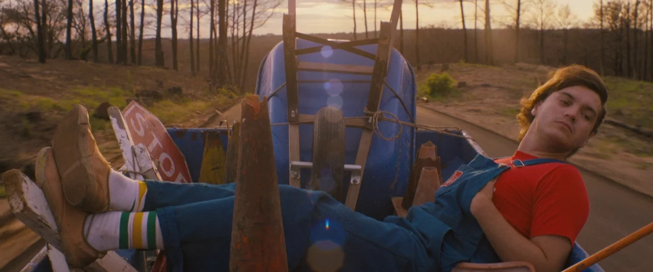 Prince Avalanche (2013) movie still: medium — A man lies in the blue metal bed of a moving truck at sunset. Cluttered construction items surround…