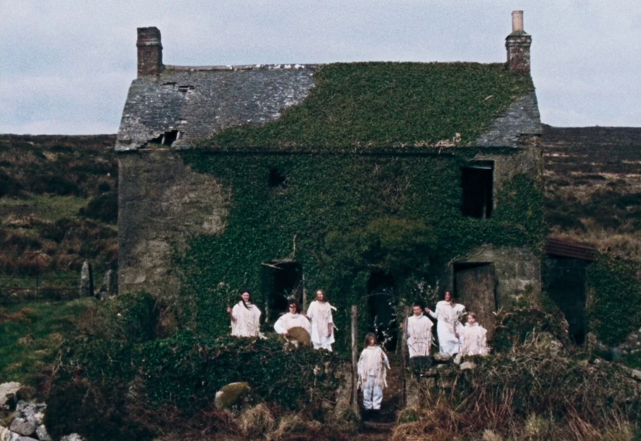 Enys Men (2022) movie still: wide — A stone cottage covered in thick green vines sits on a desolate moorland hill. Six figures in white clothing are…