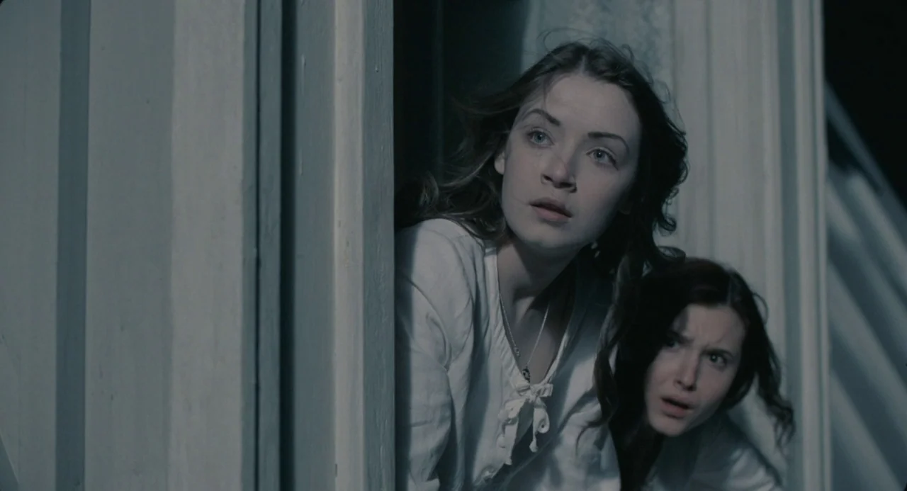 The Moth Diaries (2011) movie still: medium — Two young women peek cautiously from behind a white wooden door frame. They look toward the shadows…