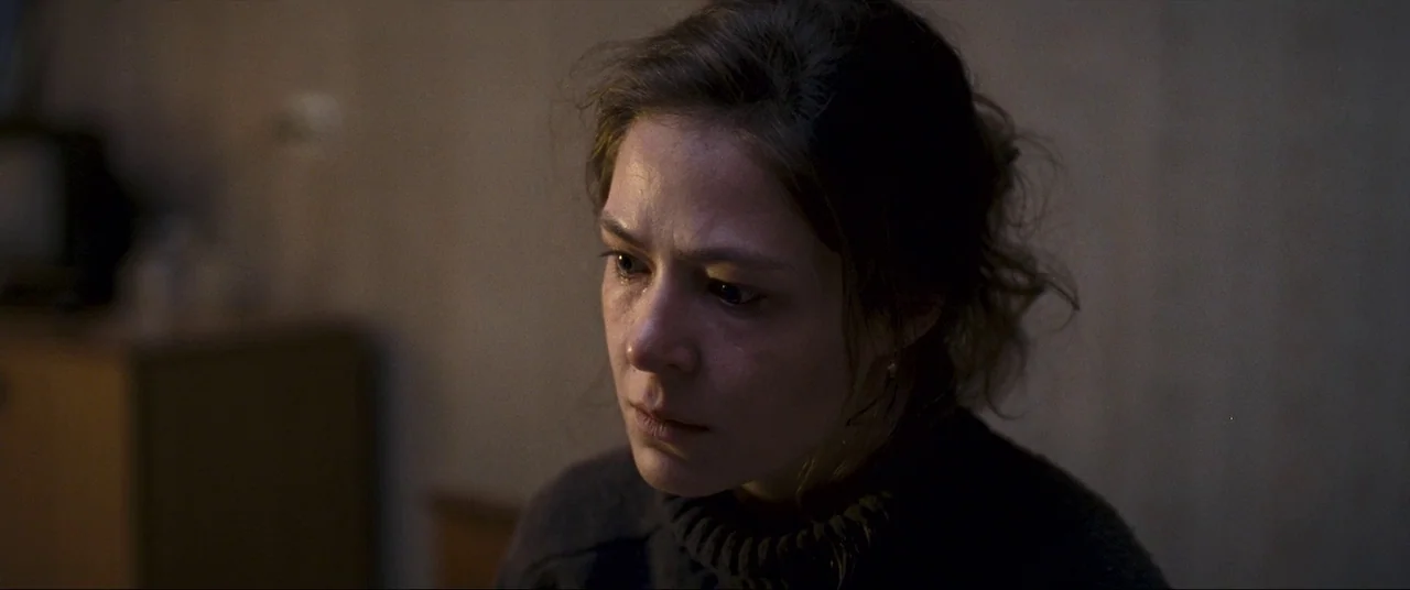 Leviathan (2014) movie still: closeup — A dimly lit room with muted-colored, nondescript walls in the background. A young woman with her hair pulled…