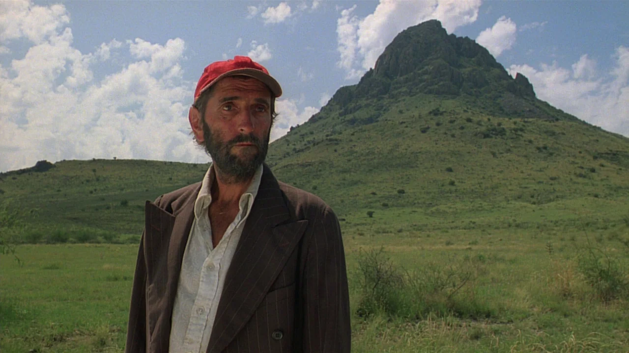 Paris, Texas (1984) movie still: medium — A grassy open plain sits before a large, solitary rocky mountain under a blue sky. A man stands in the…