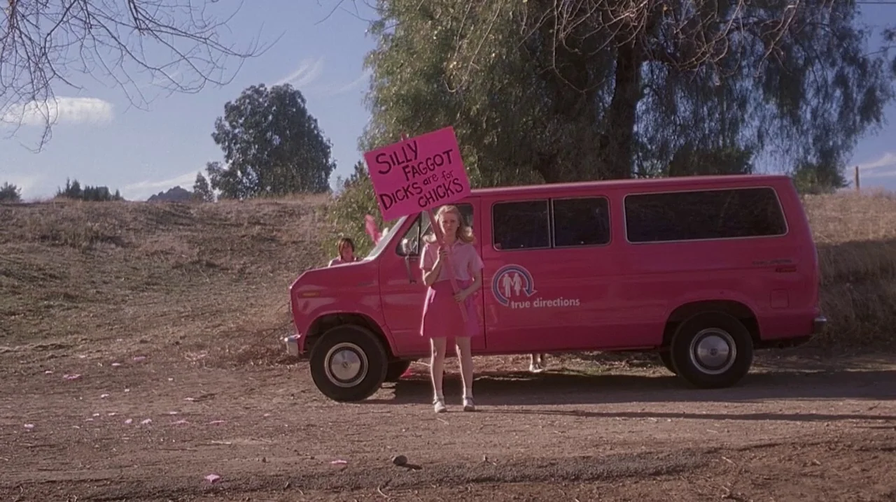 But I’m A Cheerleader (1999) movie still: fullbody — A dry, dusty dirt lot features a bright pink van with stylized branding decals. A young woman…