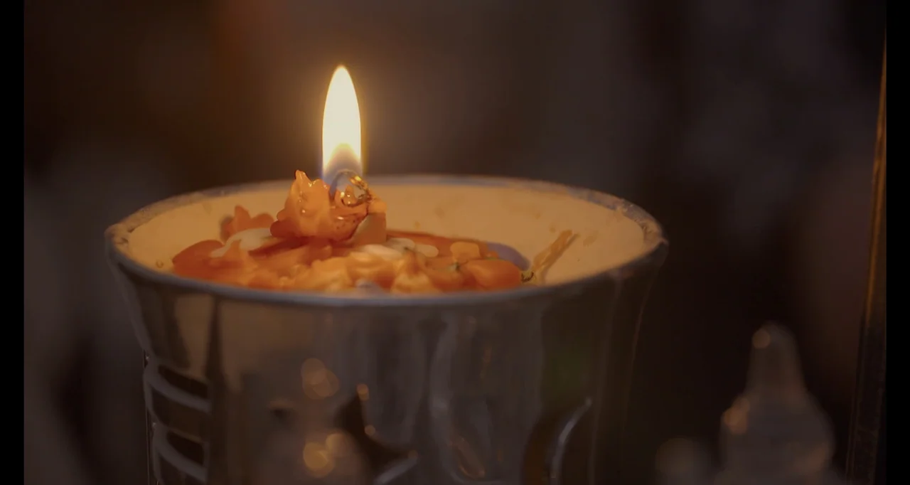 Saudi Women's Driving School (2019) movie still: closeup — A lit candle sits inside a silver metallic holder on a dark surface. The small flame…