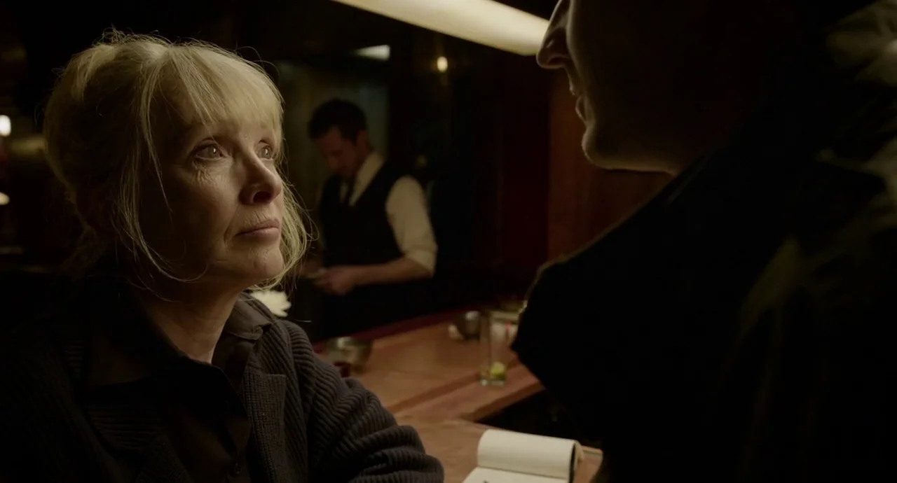 Birdman (2014) movie still: medium — A dimly lit counter interior with a wooden bar and a blurred background figure. A middle-aged woman sits at the…