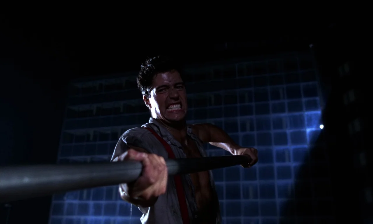Demons 2 (1986) movie still: medium — A dark rooftop is visible at night against the illuminated grid windows of a building. A man in an open shirt…