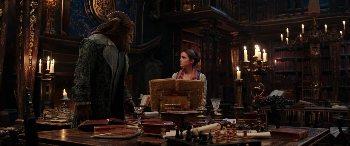 Beauty and the Beast (2017) movie still: medium — A dark, ornate library filled with towering bookshelves and wooden furniture lit by candles. The…