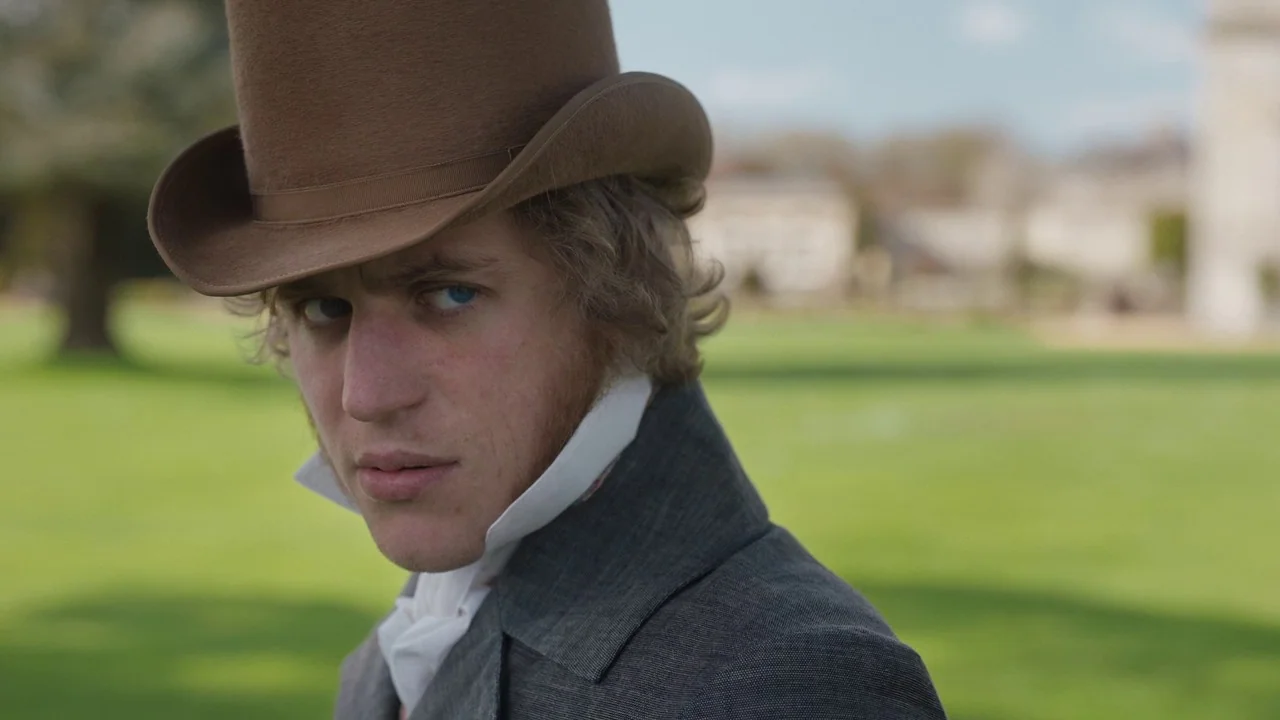 Emma. (2020) movie still: medium — A man stands outdoors on a green lawn in front of a stone building. He wears a brown top hat and a grey coat…