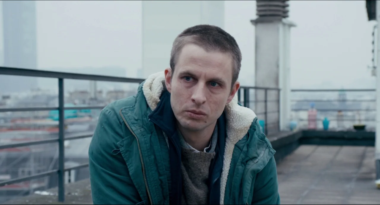 The Night Eats the World (2018) movie still: medium — A flat concrete urban rooftop overlooks a hazy, dense city skyline. A man sits quietly…