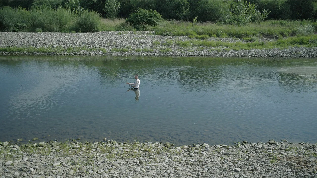 The Zone Of Interest (2023) movie still: wide — A shallow river flows past a pebbled shoreline under a clear sky. A man in a white tank top stands…