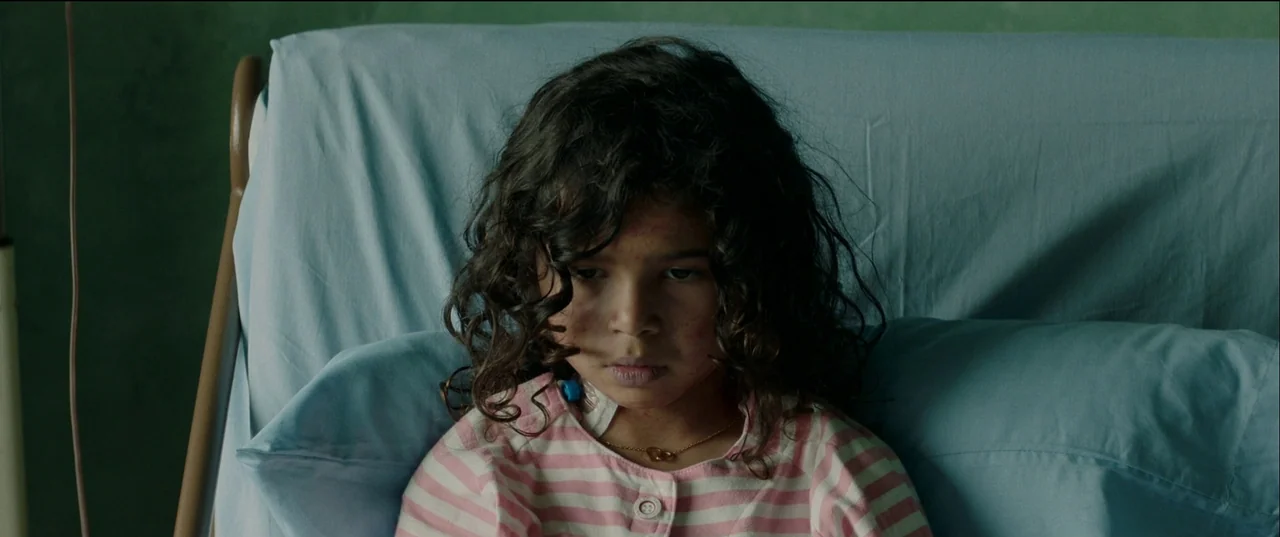 Birth/Rebirth (2023) movie still: closeup — A medical bed with light blue sheets is positioned against a muted green wall. A young child with curly…