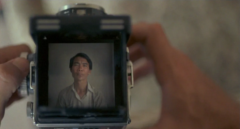 The Killing Fields (1984) movie still: closeup — A close-up view through the waist-level viewfinder of a vintage twin-lens reflex camera. A man is…