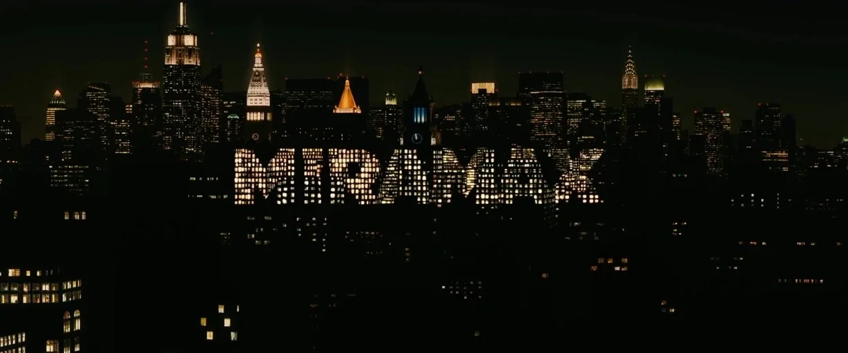The Debt (2010) movie still: establishing — A dense urban night skyline features the illuminated white letters of a film logo. The letters appear as…
