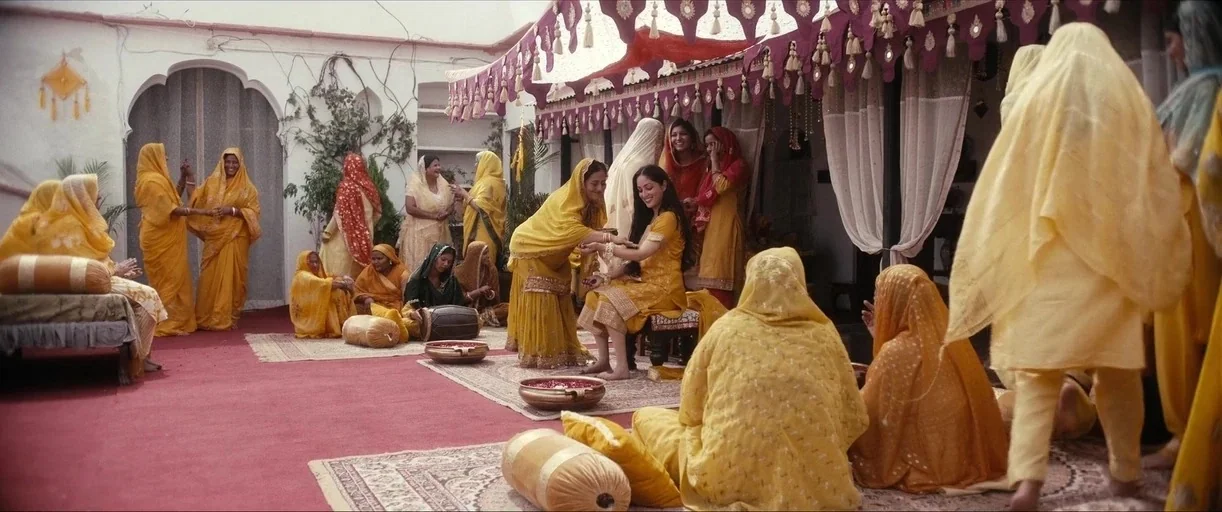 Haq (2025) movie still: wide — A sunlit outdoor courtyard features ornate fabric decorations and a red carpet. A group of women wearing yellow…