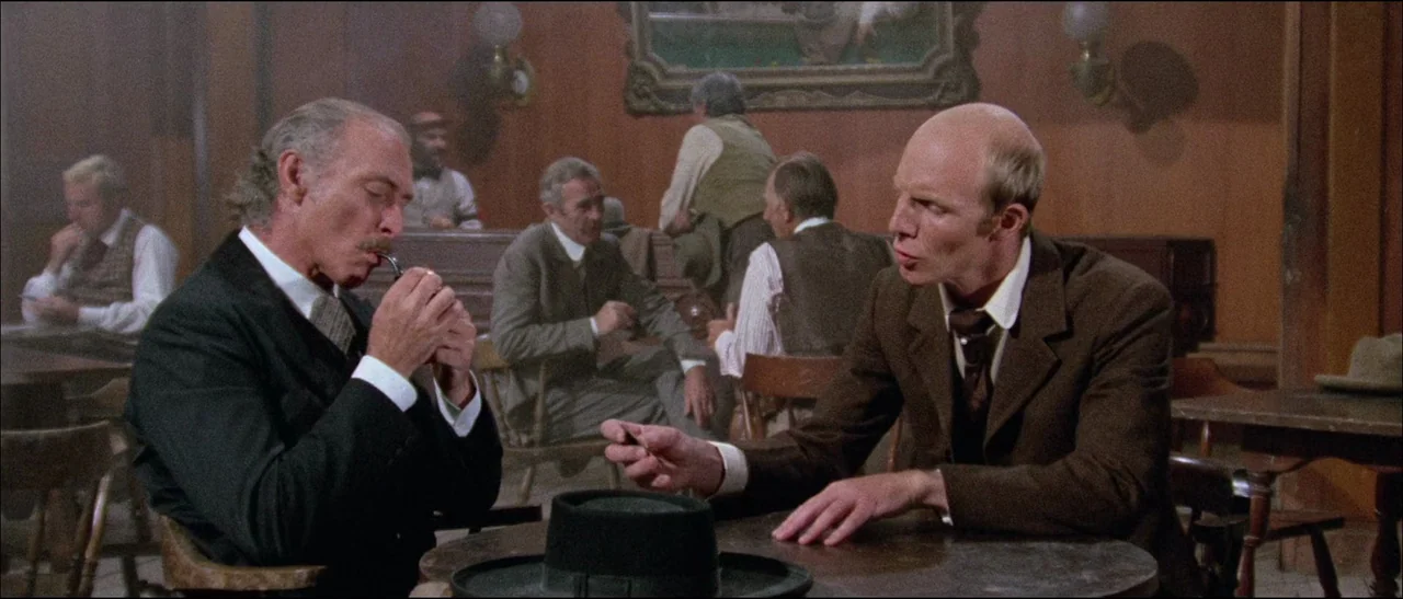 The Grand Duel (1972) movie still: medium — A dimly lit wooden saloon features period patrons sitting at tables in the background. Two men sit at a…