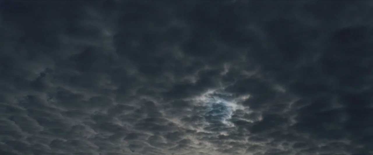 For Ellen (2012) movie still: establishing — An overcast sky filled with dense, dark grey cumulonimbus clouds. A small patch of lighter blue is…