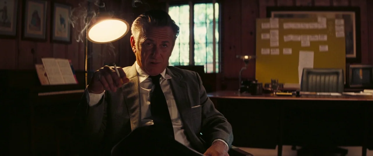 Licorice Pizza (2021) movie still: medium — A dimly lit room with wood-paneled walls holds a large wooden desk and task lamp. A middle-aged man in a…