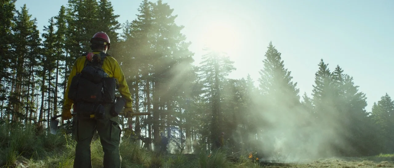 Only The Brave (2017) movie still: fullbody — A group of wildland firefighters stands in a sunlit pine forest filled with haze. One firefighter in…
