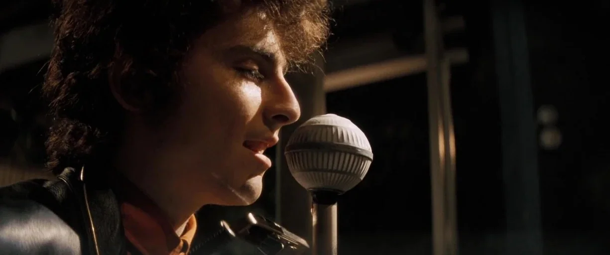 A Complete Unknown (2024) movie still: closeup — A dark, dimly lit studio environment with a prominent vintage-style microphone. A young man with…