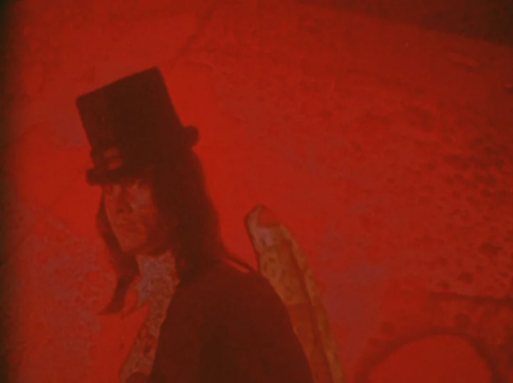 Invocation of My Demon Brother (1969) movie still: medium — A man wearing a tall top hat stands against a textured, indistinct backdrop. The scene…