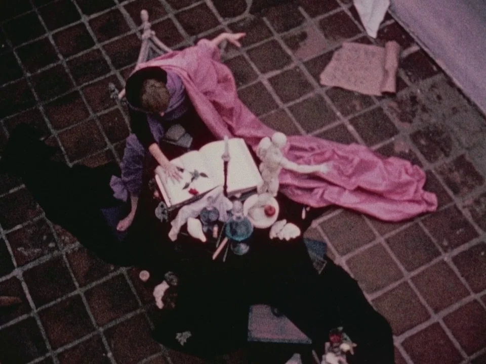 The Wormwood Star (1956) movie still: wide — A person sits on a stone tiled ground beside a small table. They reach toward an open book on the table…